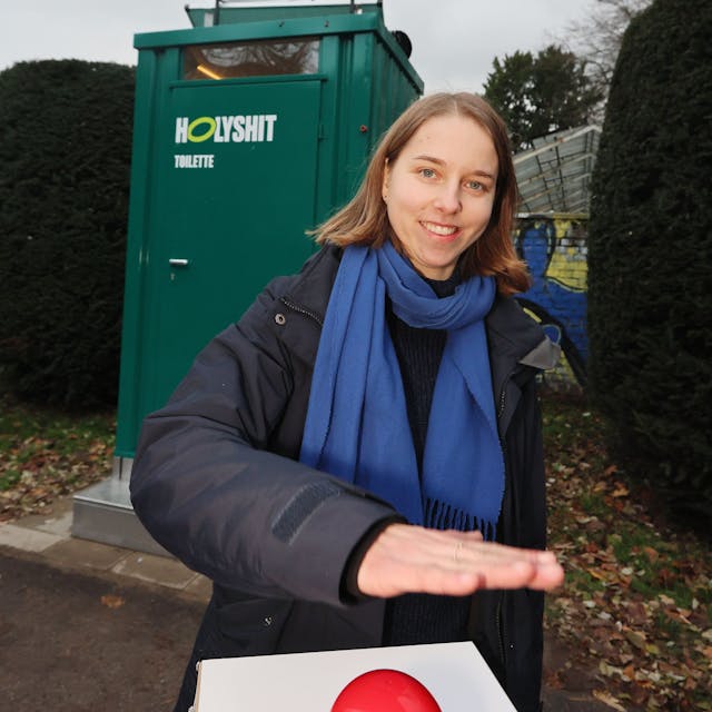 Anastasia Bondar steht vor einem grünen Toilettenhäuschen mit der Aufschrift „Holy Shit“.