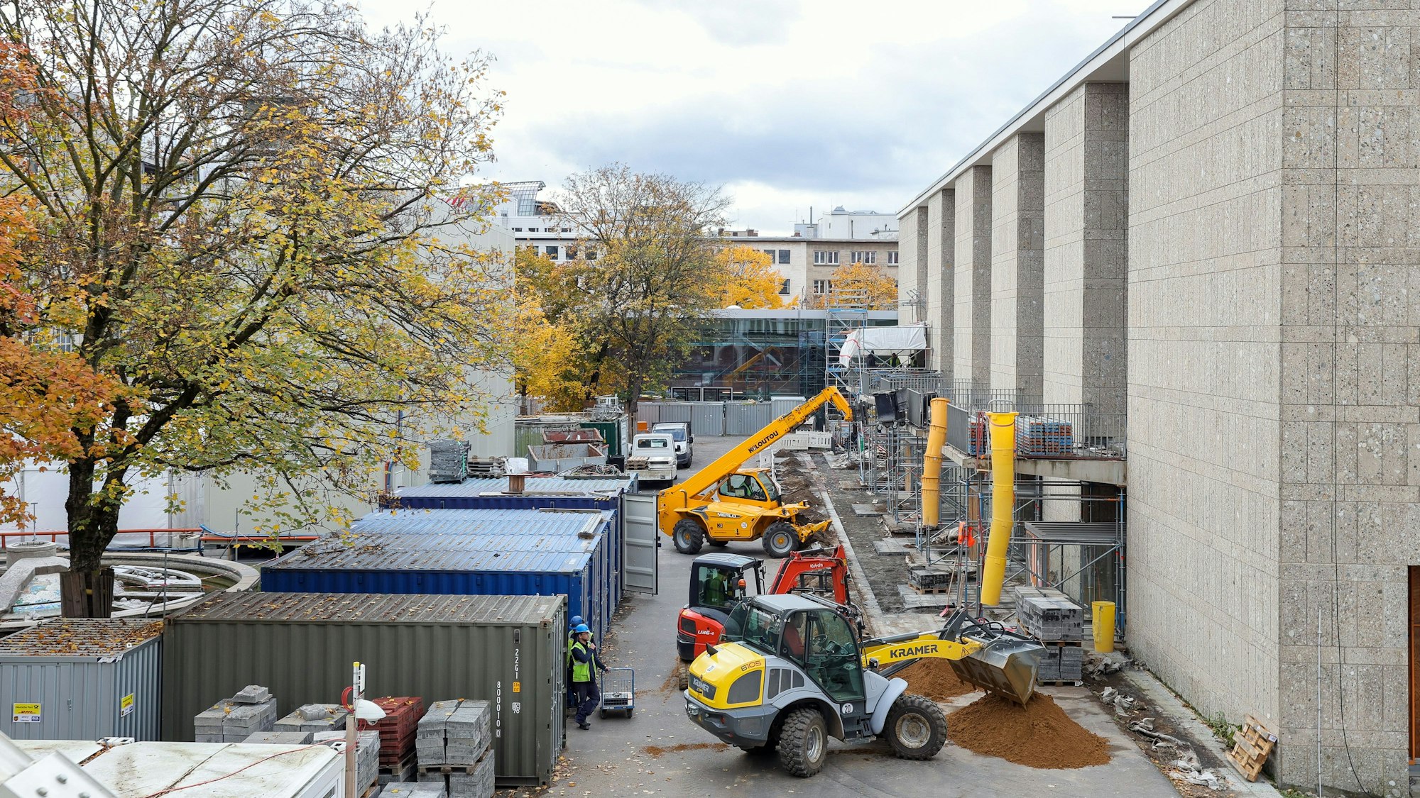 Auf dem Vorplatz der Kölner Oper verfüllen Bagger Erdreich unmittelbar vor dem Gebäude. Container, gelagertes Baumaterial sowie Gerüste am Gebäude zeugen davon, dass die Sanierungsarbeiten noch im Gange sind.