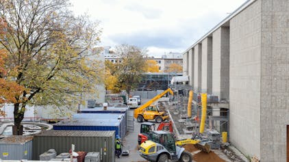 Auf dem Vorplatz der Kölner Oper verfüllen Bagger Erdreich unmittelbar vor dem Gebäude. Container, gelagertes Baumaterial sowie Gerüste am Gebäude zeugen davon, dass die Sanierungsarbeiten noch im Gange sind.&nbsp;