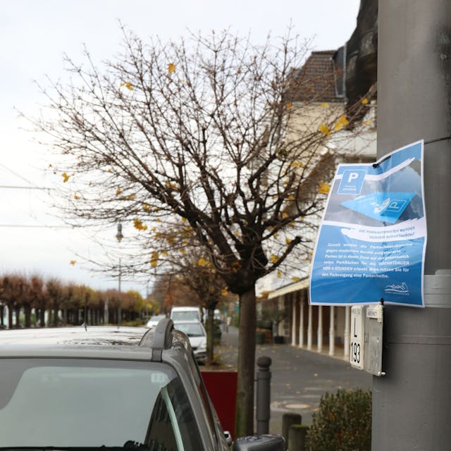 An einem Laternenpfahl neben parkenden Autos hängt ein laminiertes blaues Schild, auf dem die Stadt Autofahrer bittet, die Parkscheibe hinter die Windschutzscheibe zu legen.