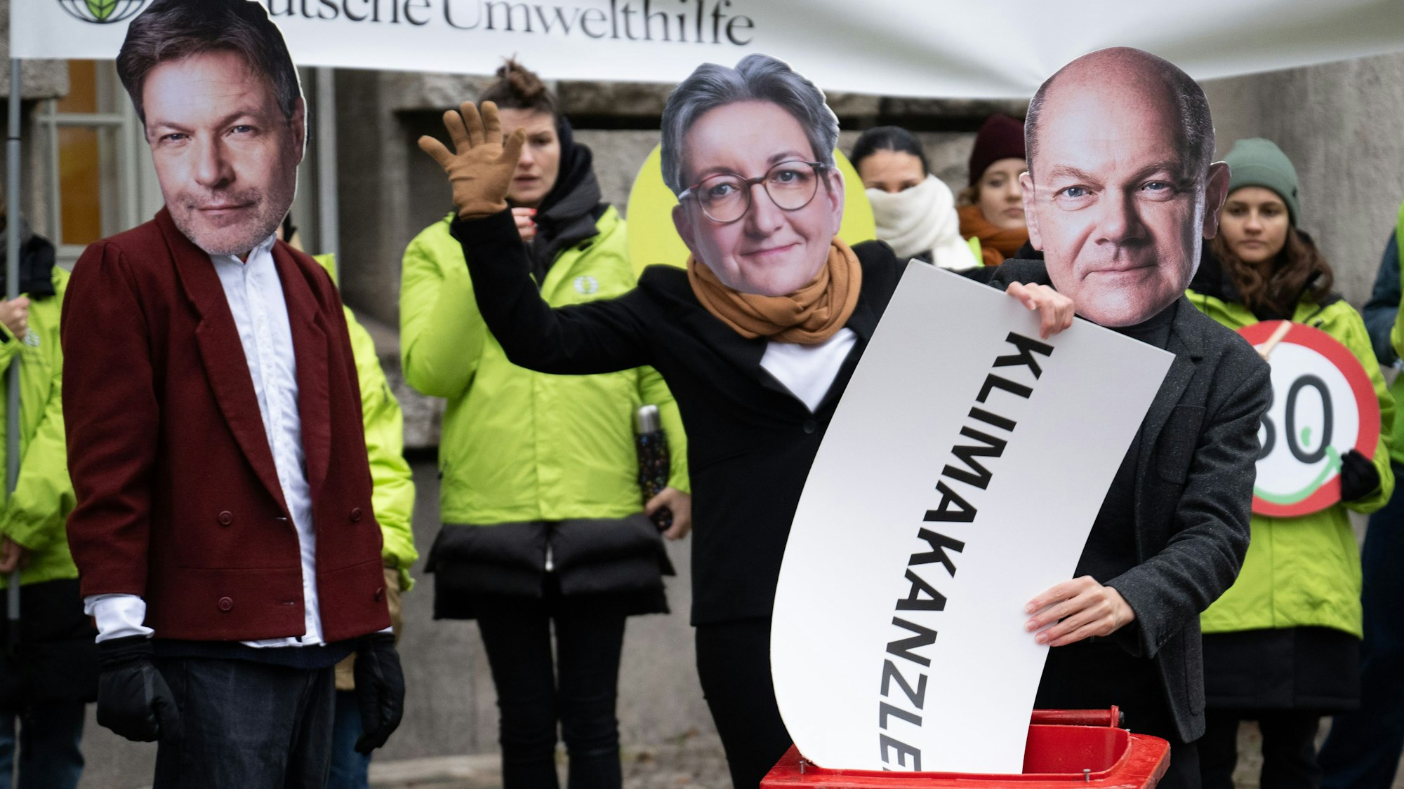 Eine Person mit einer Maske von Bundeskanzler Scholz wirft neben Aktivistinnen und Aktivisten mit Masken von Wirtschaftsminister Habeck und Bauministerin Geywitz, ein Schild mit der Aufschrift „Klimakanzler“ in eine Mülltonne.