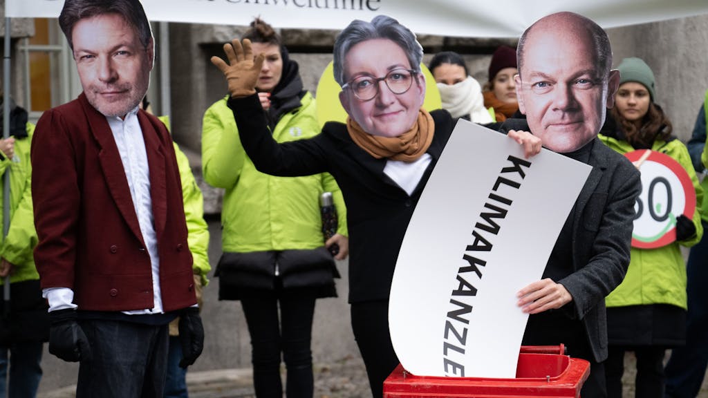 Eine Person mit einer Maske von Bundeskanzler Scholz wirft neben Aktivistinnen und Aktivisten mit Masken von Wirtschaftsminister Habeck und Bauministerin Geywitz, ein Schild mit der Aufschrift „Klimakanzler“ in eine Mülltonne.