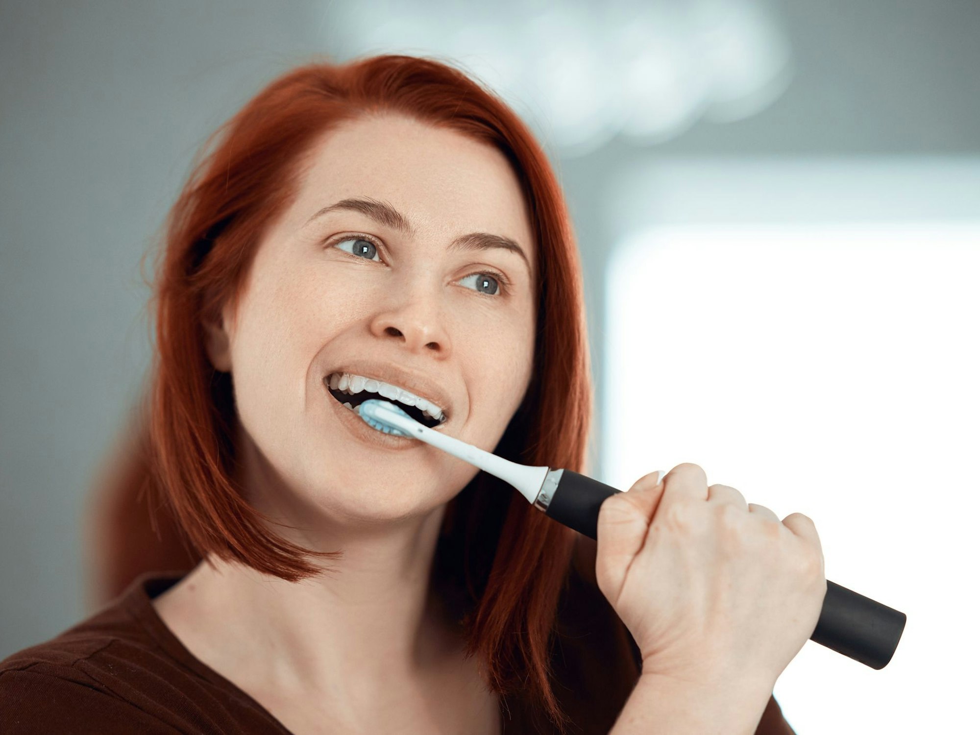 Auf dem Foto putzt sich eine rothaarige Frau die Zähne mit einer elektrischen Zahnbürste.
