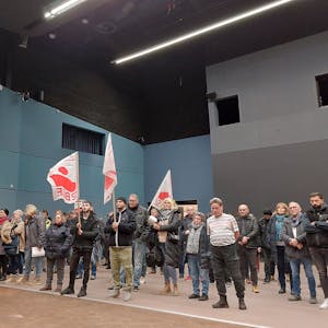 Männer und Frauen stehen in einer Veranstaltungshalle. Einige haben weiß-rote Fahnen ihrer Gewerkschaft dabei. Die Stimmung ist ernst.