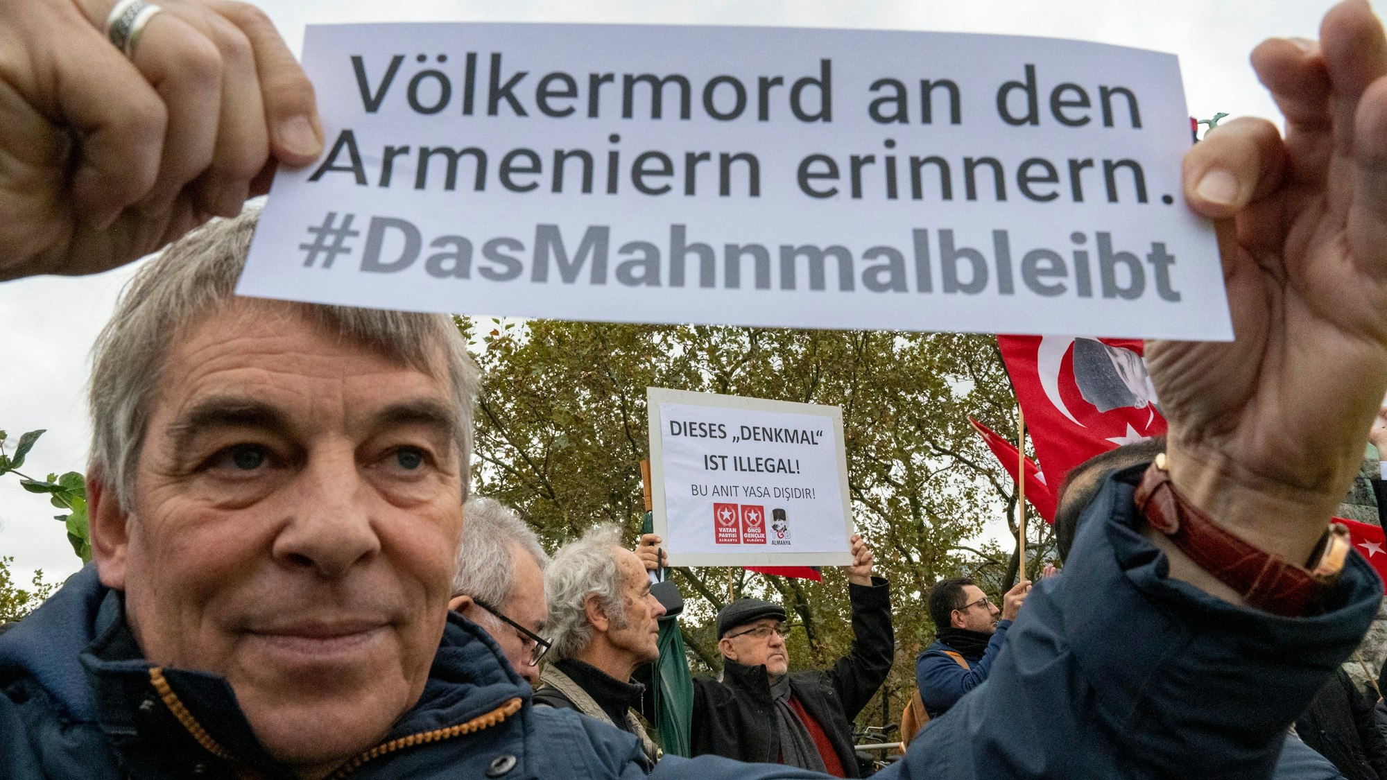 Ein Mann hält ein Schild in den Händen, darauf steht „Völkermord an den Armeniern erinnern“, im Hintergrund hält ein anderer Mann ein Schild mit der Aufschrift „Dieses ‚Denkmal‘ ist illegal“ hoch.