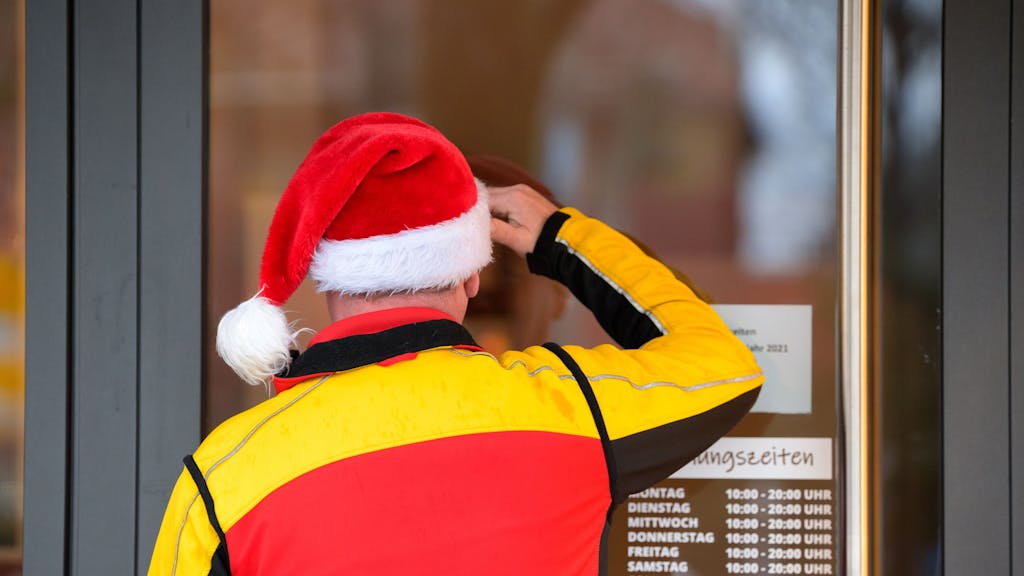 Ein Paketbote des Paketdienstes DHL mit einer Weihnachtsmütze auf dem Kopf schaut suchend durch die Eingangstür eines Ladengeschäfts.