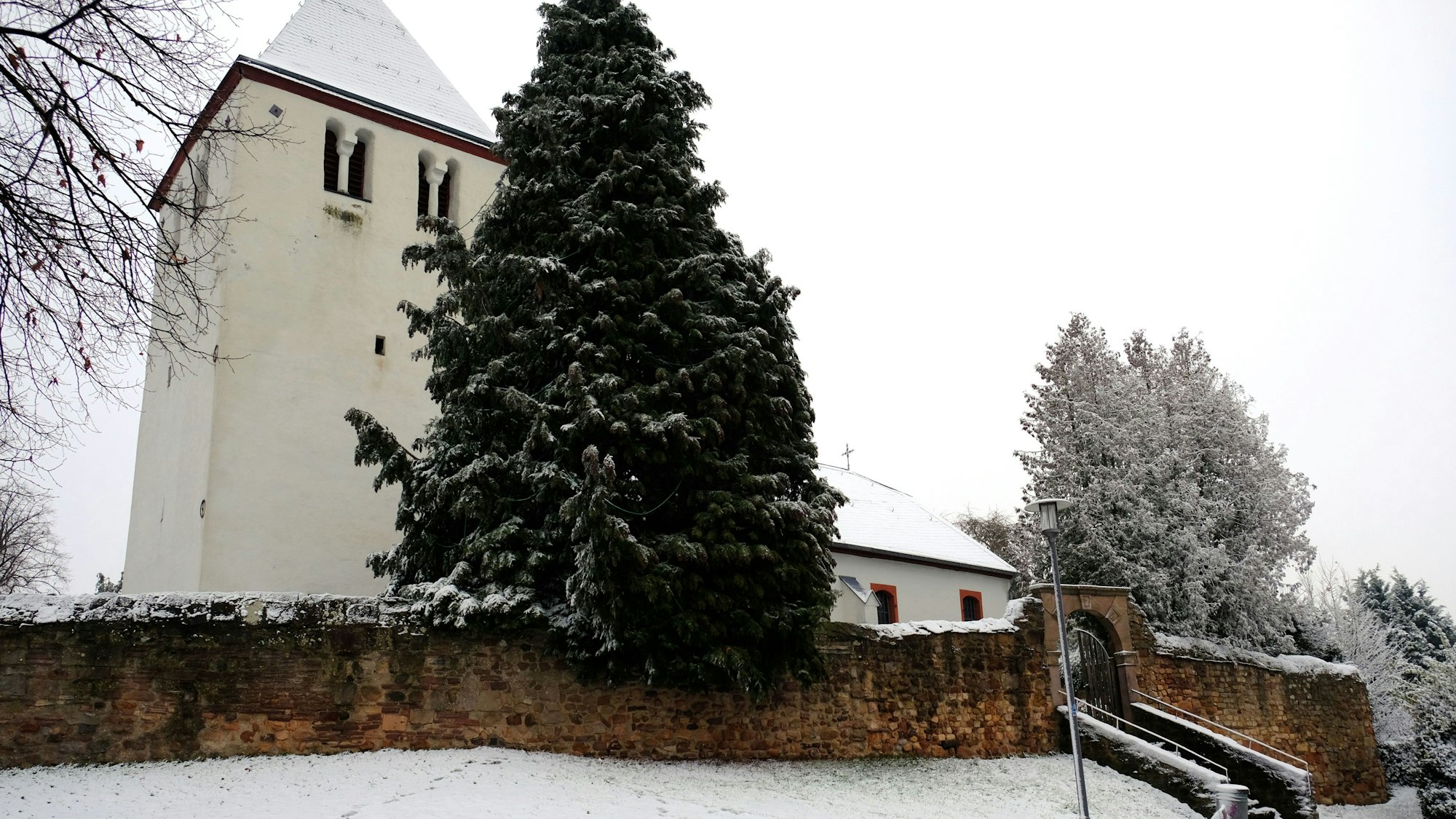 Zwischen verschneiten Bäumen und hinter einer alten Mauer ist die Alte Kirche Mechernich zu sehen.
