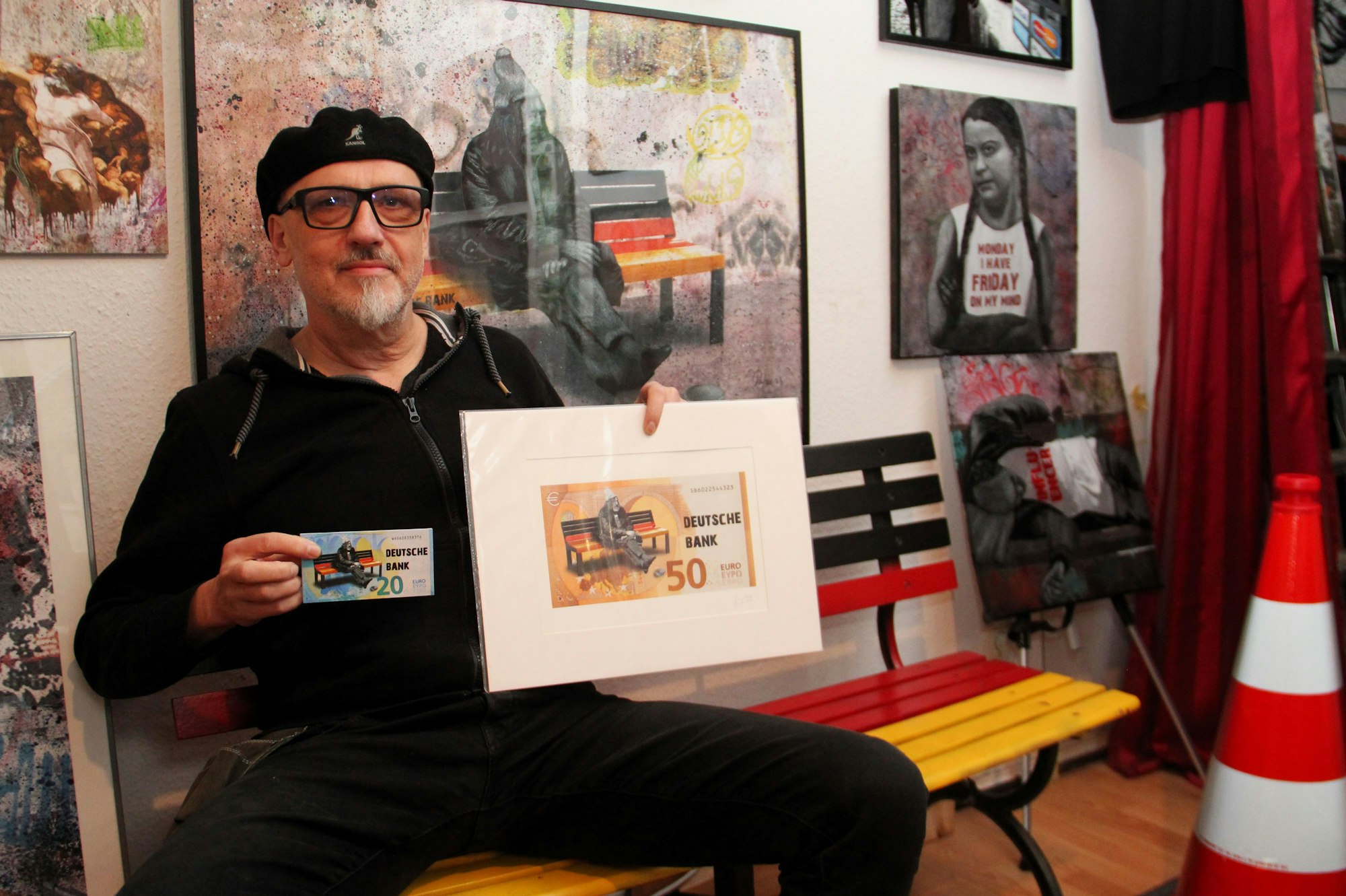 Der Künstler Peter Mück hält seine Werke in der Hand: Abbilder eines 20-Euro-Scheins und eines 50-Euro-Scheins. Darauf ist das Motiv eines Mannes, der auf einer schwarz-rot-golden lackierten Bank sitzt, zu erkennen.