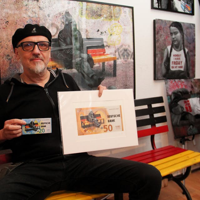 Der Künstler Peter Mück hält seine Werke in der Hand: Abbilder eines 20-Euro-Scheins und eines 50-Euro-Scheins. Darauf ist das Motiv eines Mannes, der auf einer schwarz-rot-golden lackierten Bank sitzt, zu erkennen.&nbsp;