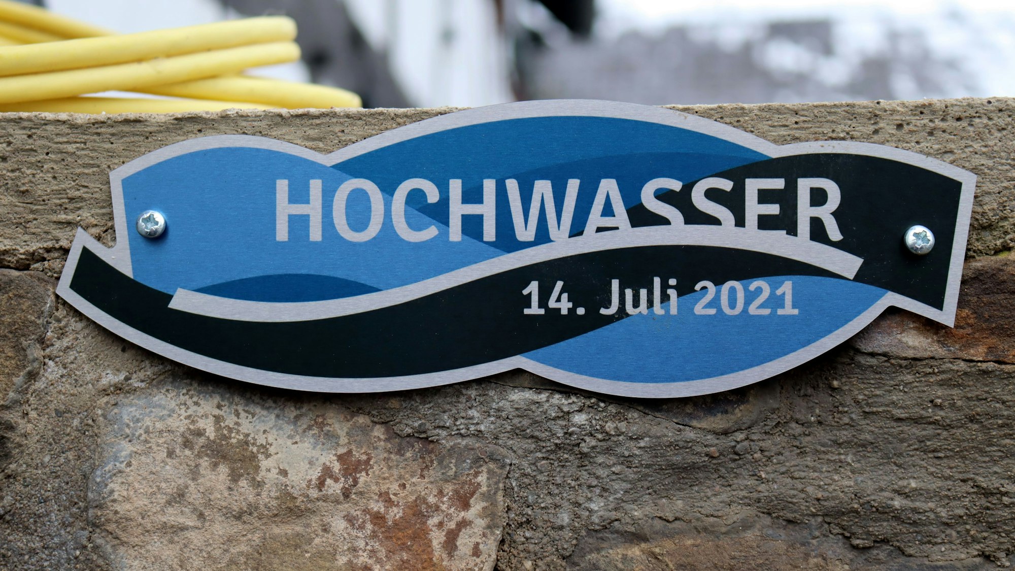 Das Bild zeigt eine Flutmarke. Sie ist in Bau und Schwarz gehalten und trägt die Aufschrift „Hochwasser 14. Juli 2021“.