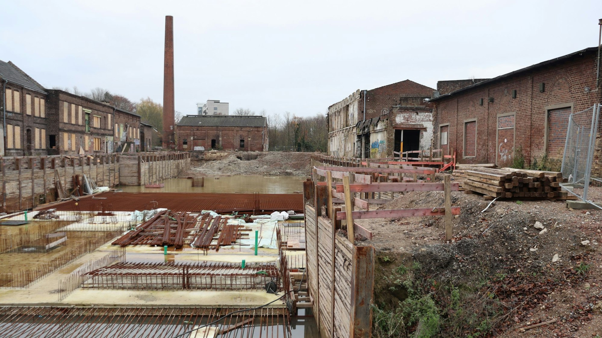 Eine Baugrube mit einem alten Industrie-Schornstein im Hintergrund ist zu sehen.