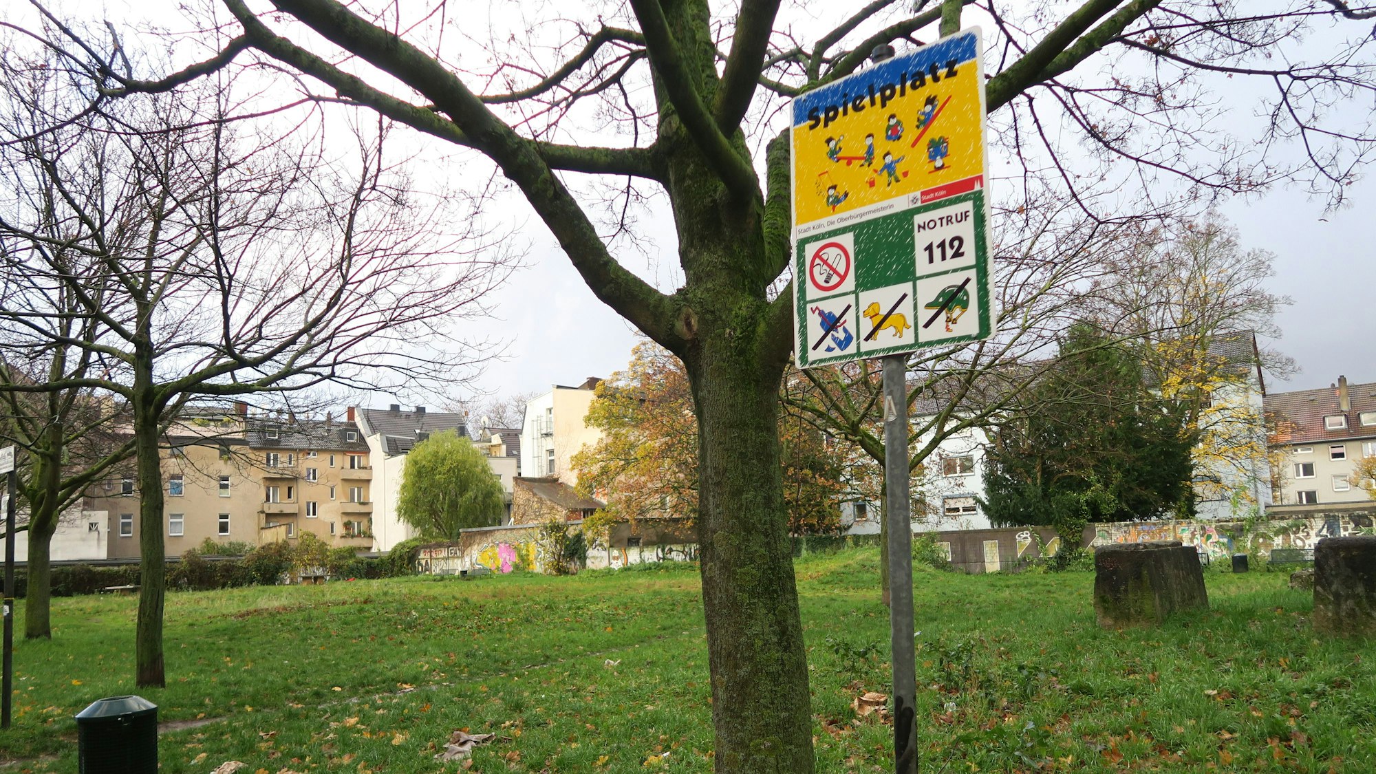 Eine Wiese mit Bäumen und einem Spielplatz-Schild ist zu sehen, im Hintergrund sind Wohnhäuser.