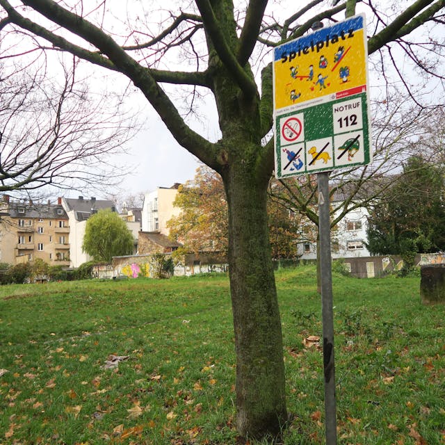 Eine Wiese mit Bäumen und einem Spielplatz-Schild ist zu sehen, im Hintergrund sind Wohnhäuser.&nbsp;