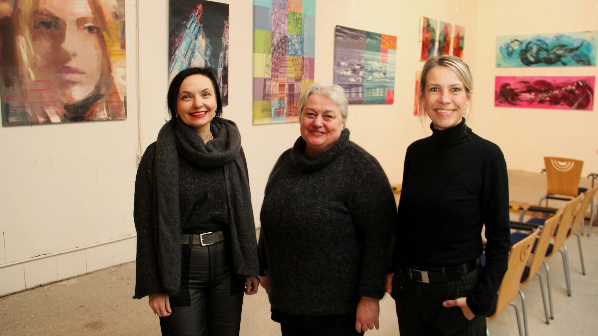 Drei schwarz gekleidete Frauen stehen nebeneinander vor einer Wand, an der Bilder hängen.