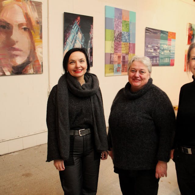 Drei schwarz gekleidete Frauen stehen nebeneinander vor einer Wand, an der Bilder hängen.