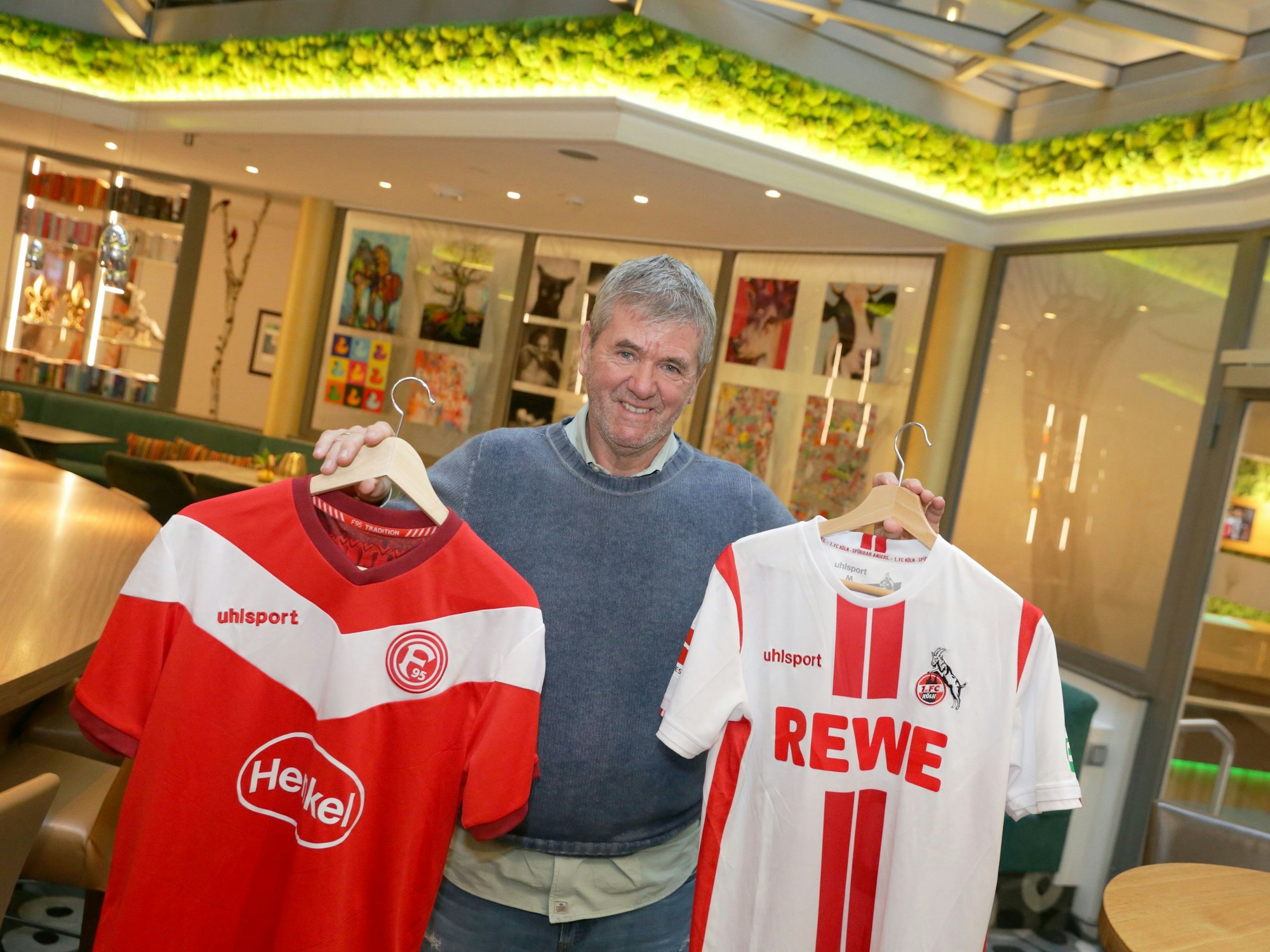 Friedhelm Funkel hält zwei Trikots von Fortuna Düsseldorf und dem 1. FC Köln in die Kamera.