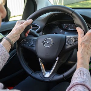 Das Symbolbild zeigt eine Seniorin, die am Steuer eines Autos sitzt. Sie umfasst mit beiden Händen das Lenkrad.