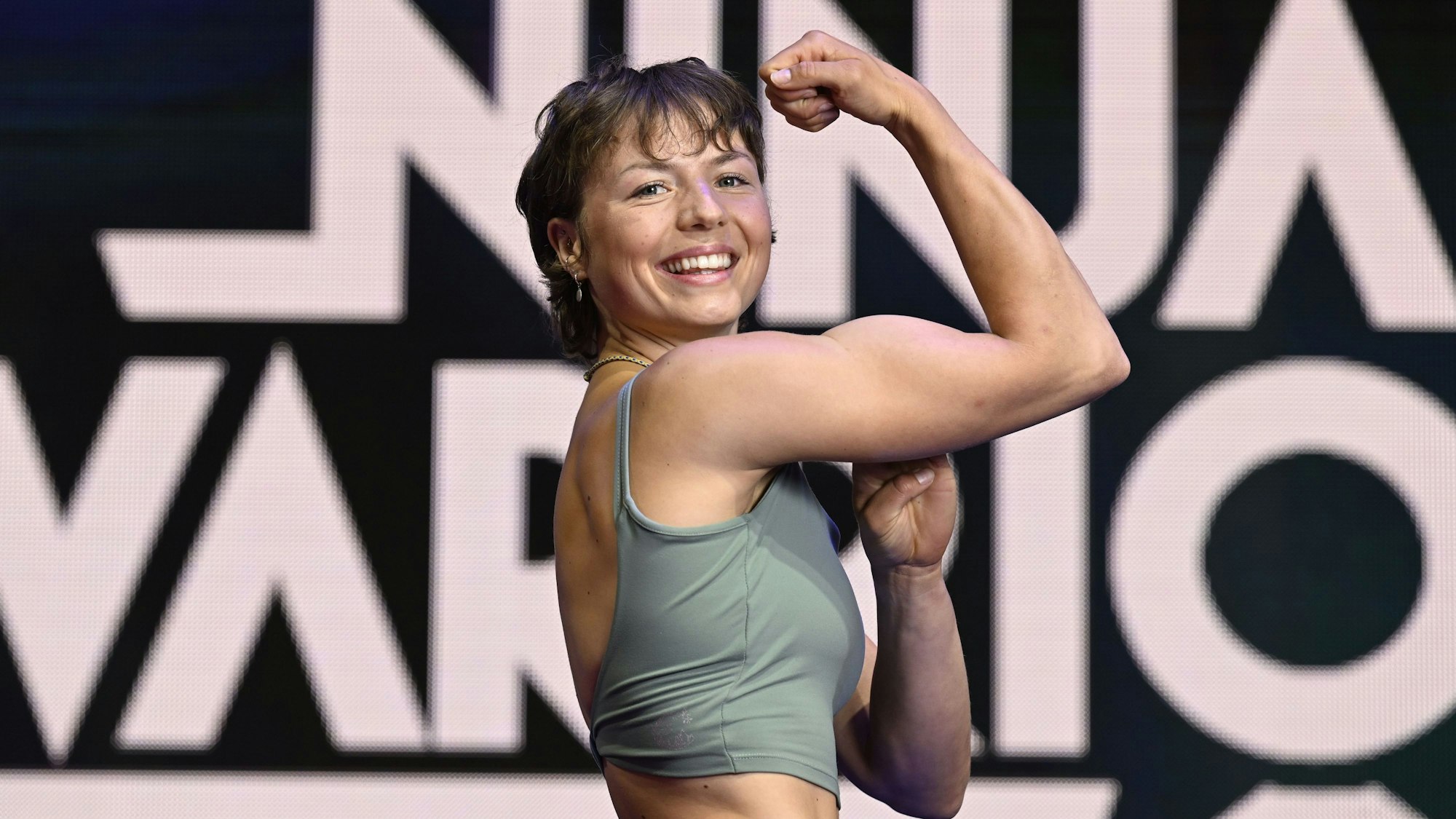 Sportstudentin Ada Theilken zeigt vor einem Schriftzug der RTL-Show Ninja Warrior Germany ihre Muskeln.