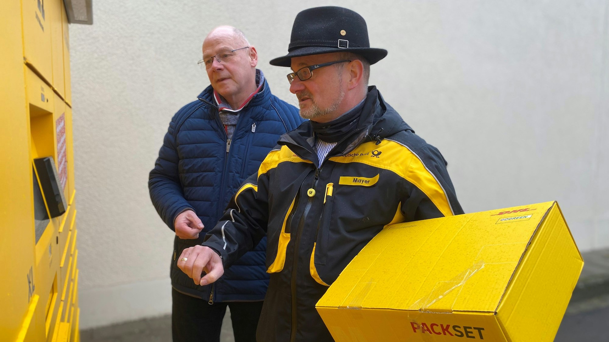 Alexander Eskes, Erster Beigeordneter der Gemeinde Weilerswist und Peter Mayer, der regionale Politikbeauftragte der Deutschen Post, stehen vor der Poststation 711 an der Meckenheimer Straße 40 in Weilerswist. Mayer hält ein Paket in den Händen.