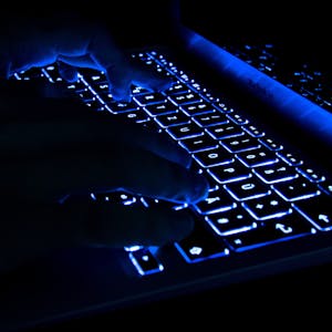 Ein Mann tippt in der Dunkelheit auf erleuchtete Tasten eines Laptops.