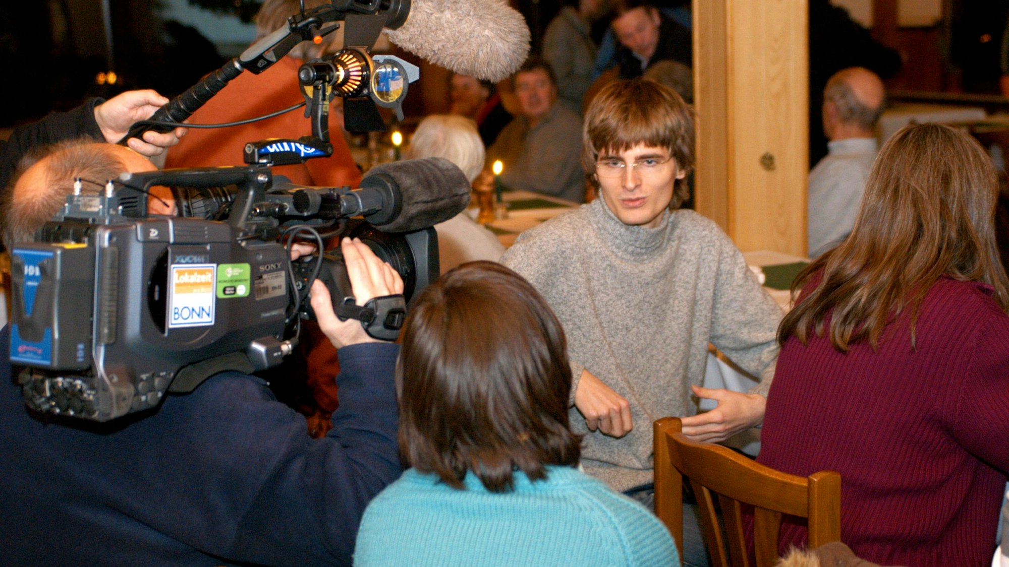In einer Versammlung sitzt der damals 24-jährige Arvid Bell auf einem Stuhl. Ein Fernsehteam mit Kamera und Mikrofon nimmt ihn für ein Porträt auf.