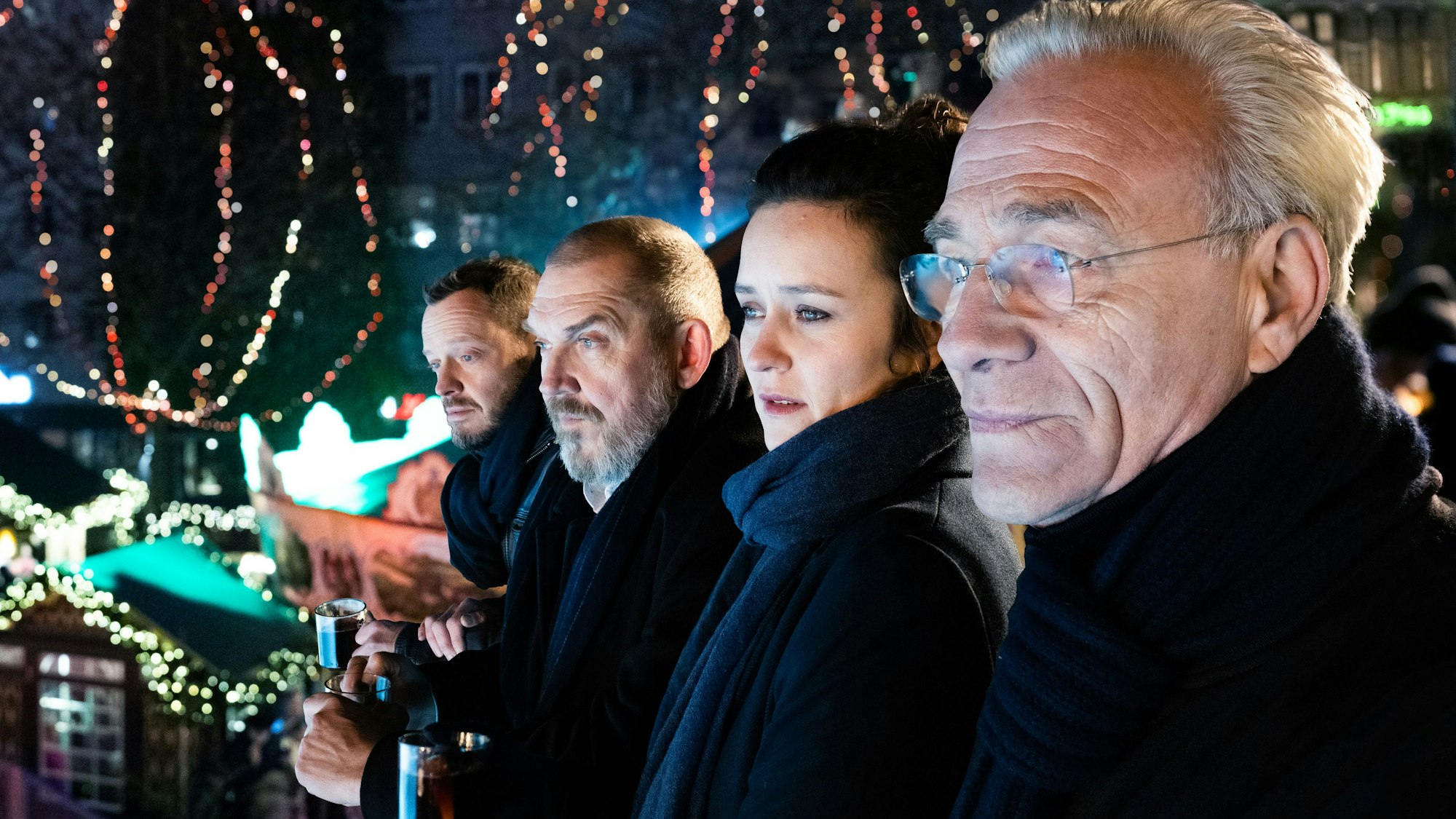 Drei Männer und eine Frau schauen alle in dieselbe Richtung, im Hintergrund sind die Lichter eines Weihnachtsmarkts zu sehen.