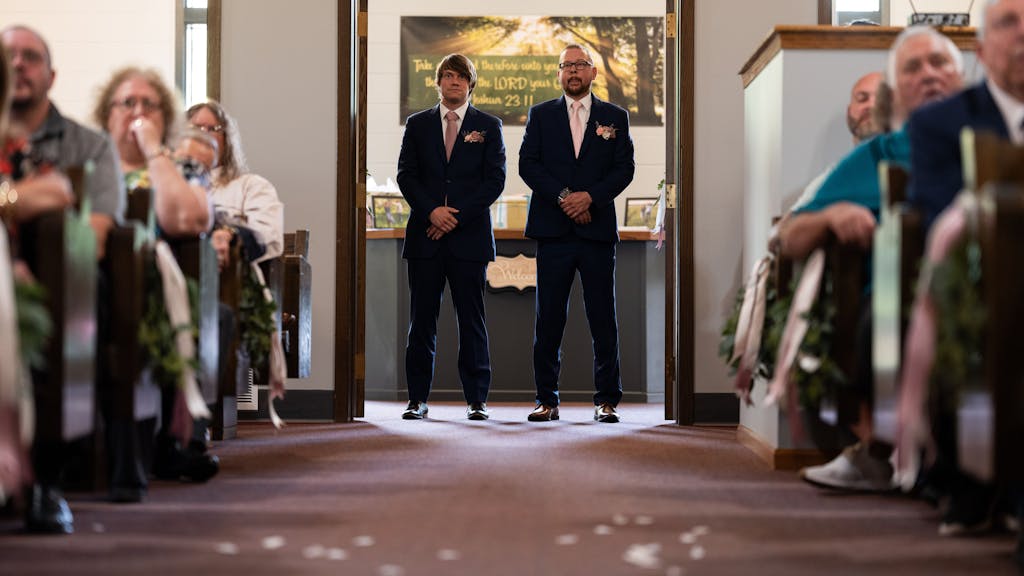 Jan Rolfes steht nehmen Ben Clark, beide in festlichen Anzügen, während die Hochzeitsgäste in den Bänken warten.
