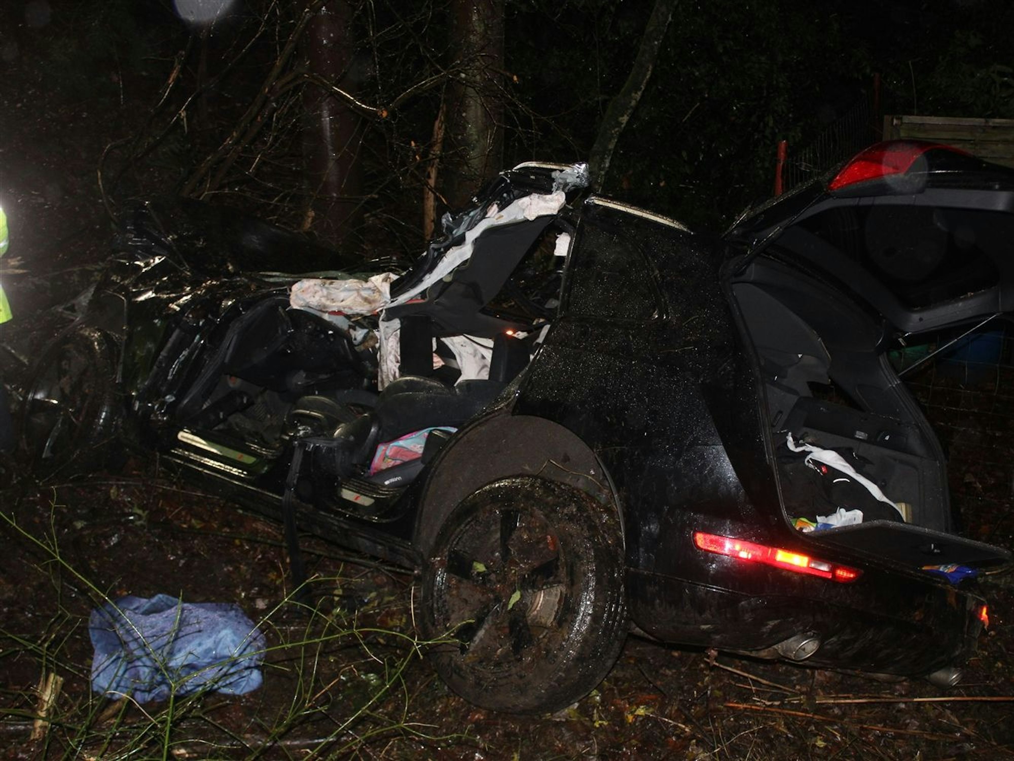 Foto eines schwarzen Autos, das nach einem Unfall zerstört in einem Waldstück liegt.