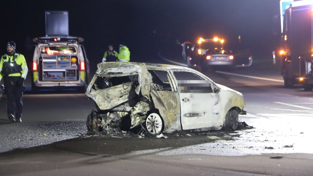 Ein Kleinwagen steht ausgebrannt und mit stark beschädigtem Heck quer auf der Autobahn, Einsatzkräfte sind vor Ort.