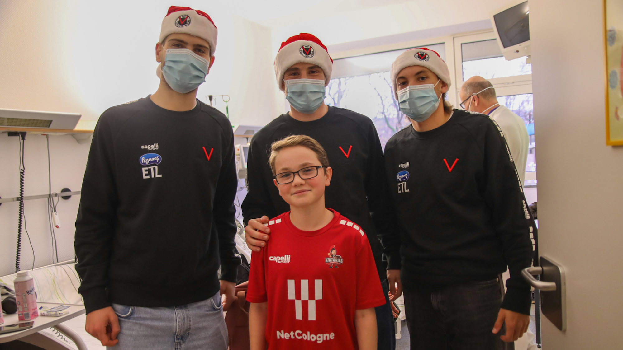 Deri junge Männer in schwarzen Pullovern, mit Weihnachtsmützen und OP-Masken und ein Junge mit Brille in einem roten T-Shirt stehenvor einem Krankenhausbett.