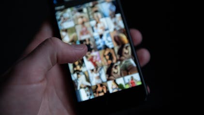 Eine Hand hält ein Handy, der Bildschirm zeigt pornografische Fotos.
