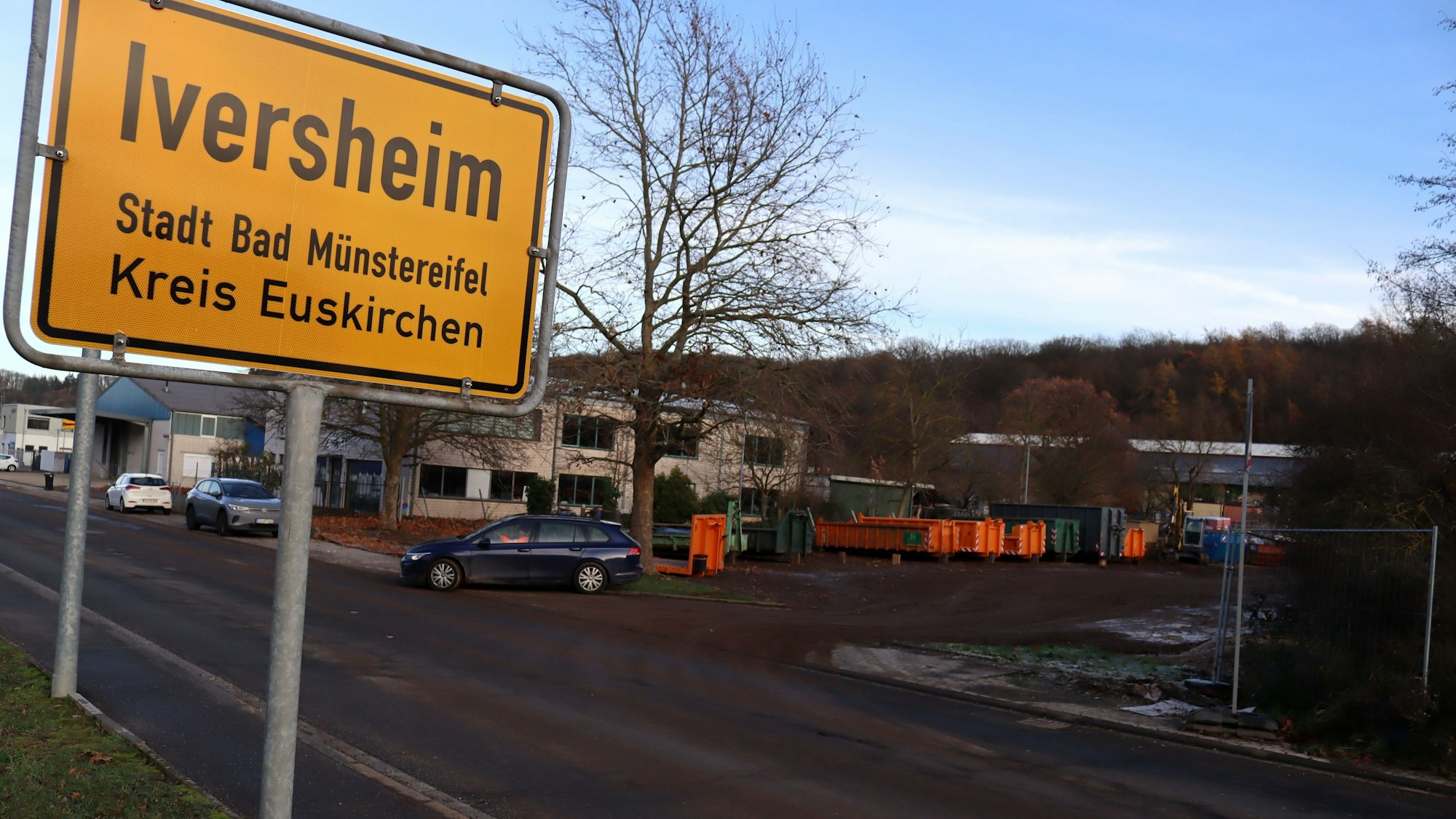 Das Ortsschild von Iversheim ist präsent, im Hintergrund ist eine mit Bauschutt-Containern besetzte Fläche zu sehen.