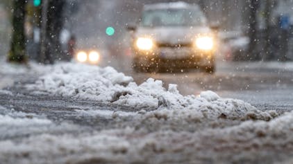 Schnee und Eis liegen am Straßenrand, während ein Auto durch den Schnee fährt. Der Deutsche Wetterdienst (DWD) erwartet zum Wochenende Neuschnee und Eisregen in Köln und der Region. Autofahrer könnten durch den gefrierenden Regen überrascht werden. (Symbolbild)