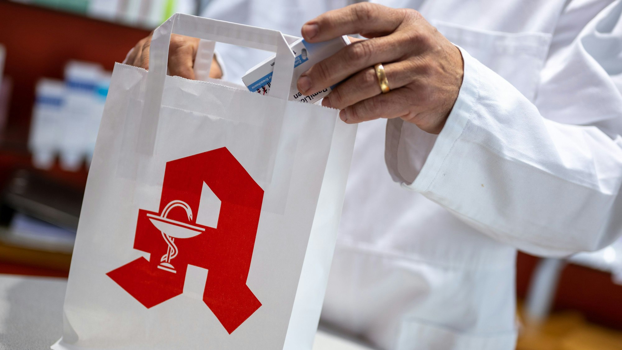 Ein Apotheker legt in einer Apotheke eine Packung mit Tabletten in eine Papiertasche mit Apotheken-Logo.