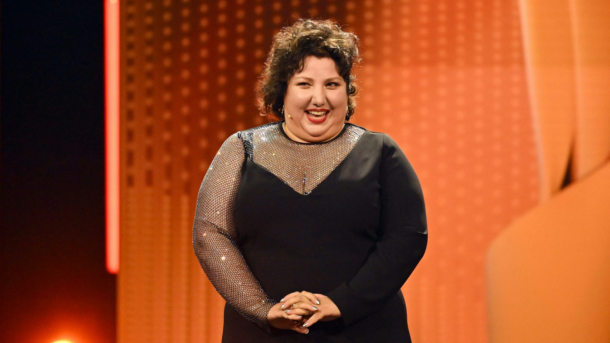 Meltem Kaptan steht in einem schwarzen Kleid auf einer Bühne, im Hintergrund sind orange Animationen zu sehen.
