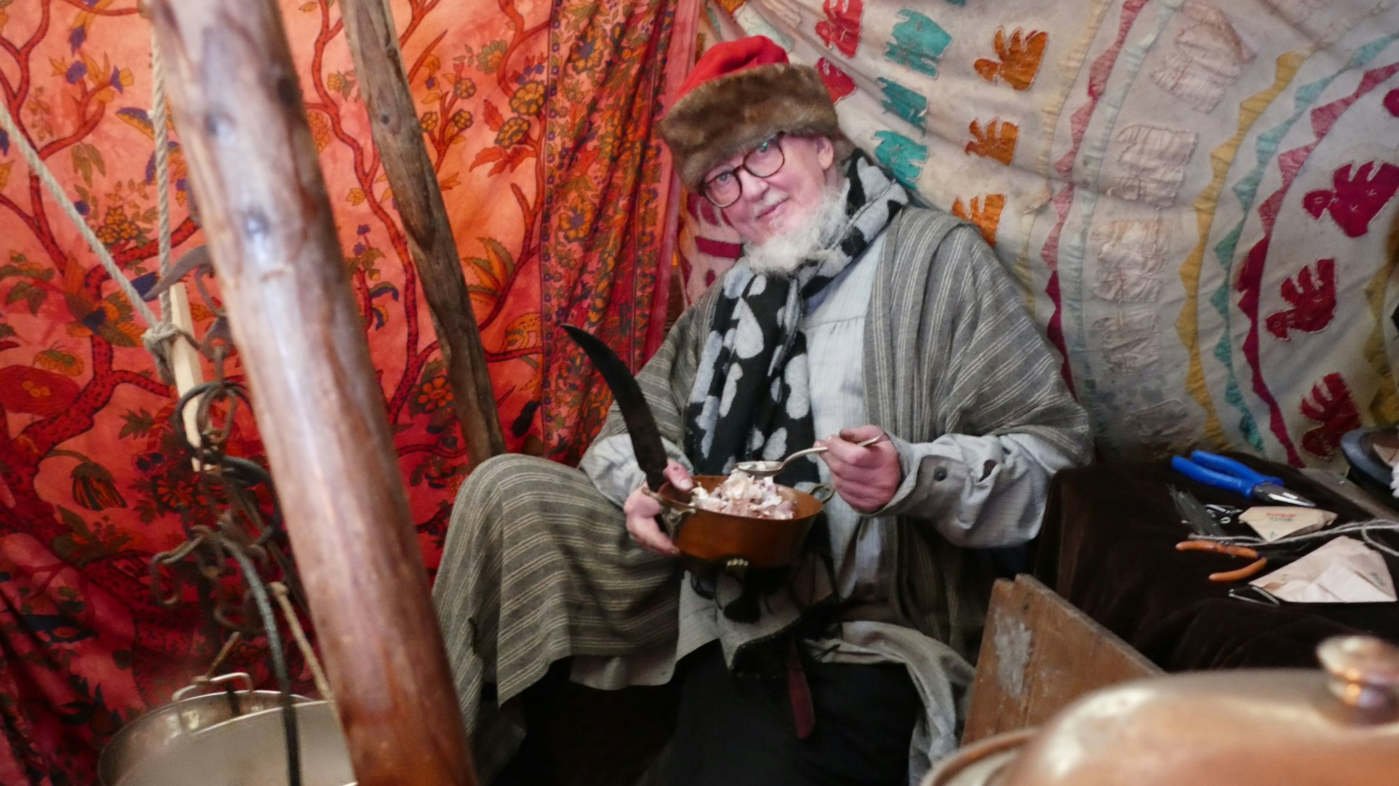 Ein Mann in mittelalterlicher Tracht sitzt in einem Zelt und löffelt Essen aus einem Topf.