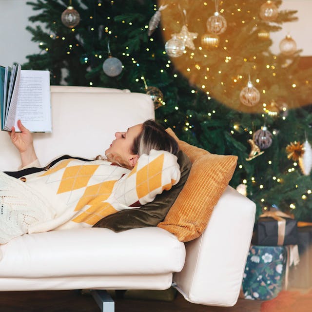 Eine Frau liest, während sie auf dem Sofa im Wohnzimmer mit geschmücktem Weihnachtsbaum liegt