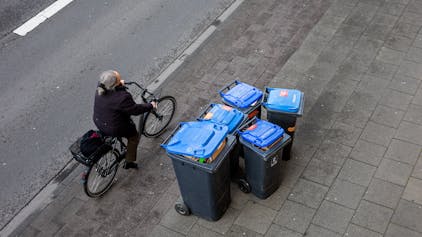 Abfalltonnen mit blauem Deckel stehen zur Abholung der Kölner Abfallwirtschaftsbetriebe am Straßenrand.