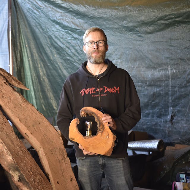 Björn Schütz steht in einem Schuppen neben alten Holzbalken. In der Hand hält er eine Lampe aus einer Baumscheibe.