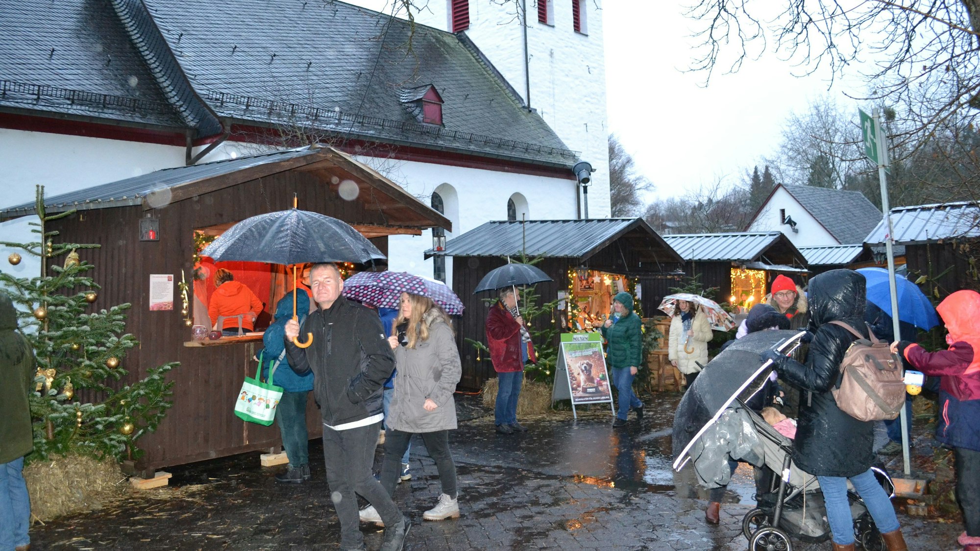 Beleuchtete Holzhütten vor einer Dorfkirche, Besucher mmit Regenschirm