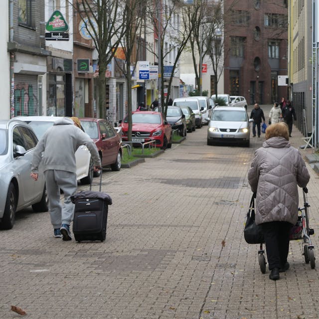 Drei Fußgänger gehen auf der Steprathstraße, eine Frau schiebt einen Rollator, ein Mann zieht einen Koffer hinter sich her. Ihnen kommt ein Auto entgegen.