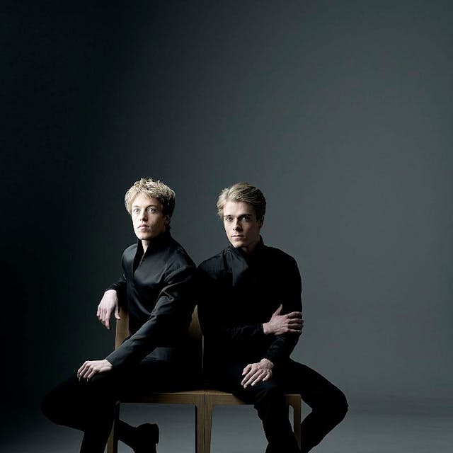 Lucas und Arthur Jussen Pianisten. Sie tragen schwarze Anzüge und sitzen auf zwei Stühlen nebeneinander. Der Hintergrund ist grau.