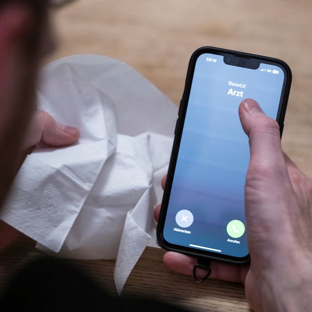 Ein Mann hält ein Taschentuch in einer Hand und ein Telefon mit dem Schriftzug ‚Arzt‘ in der Anderen