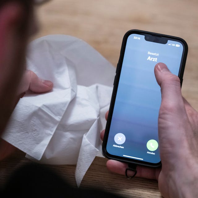 Ein Mann hält ein Taschentuch in einer Hand und ein Telefon mit dem Schriftzug ‚Arzt‘ in der Anderen
