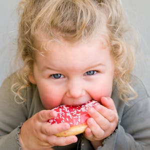 Ein kleines Mädchen isst einen pinken Donut. Symbolbild