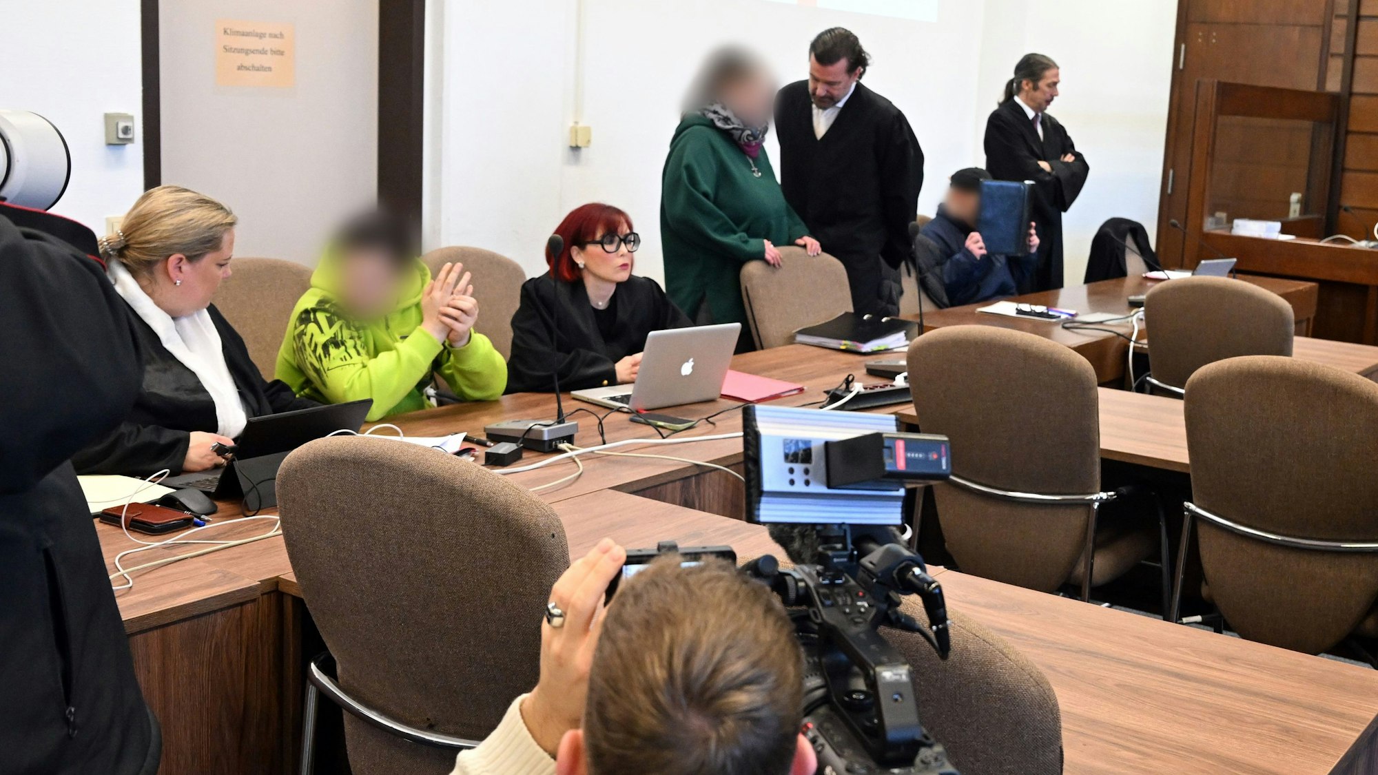 Zwei Männer sitzen auf der Anklagebank, eine Angeklagte unterhält sich stehend mit ihrem Verteidiger.