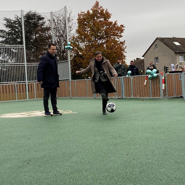 Auf einem Tartan-Platz kickt Schulleiterin Karin Bossant unter den Augen des Bürgermeisters Sacha Reichelt einen Fußball.