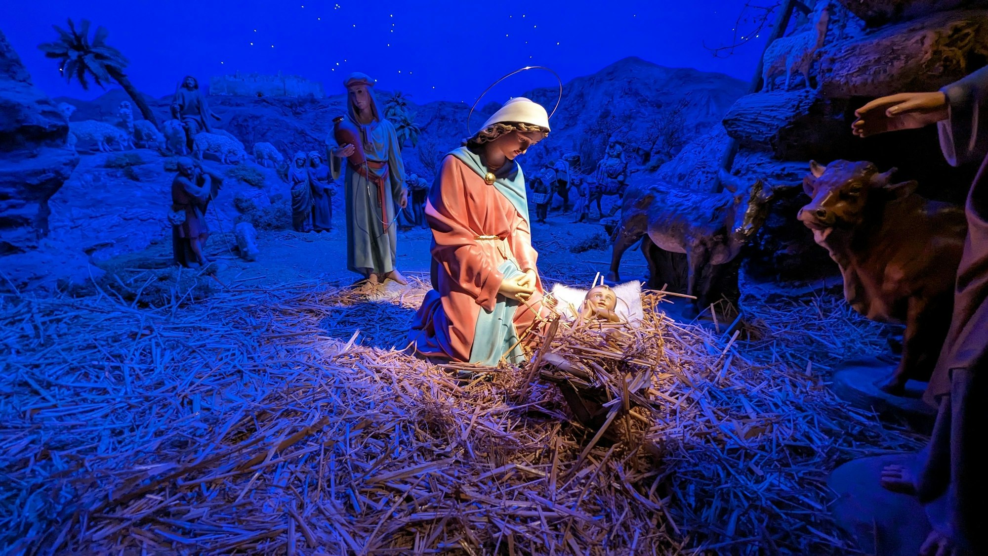 Das Bild zeigt eine Marienfigur, die vor einer mit Stroh gefüllten Krippe kniet, in der eine Babyfigur liegt.