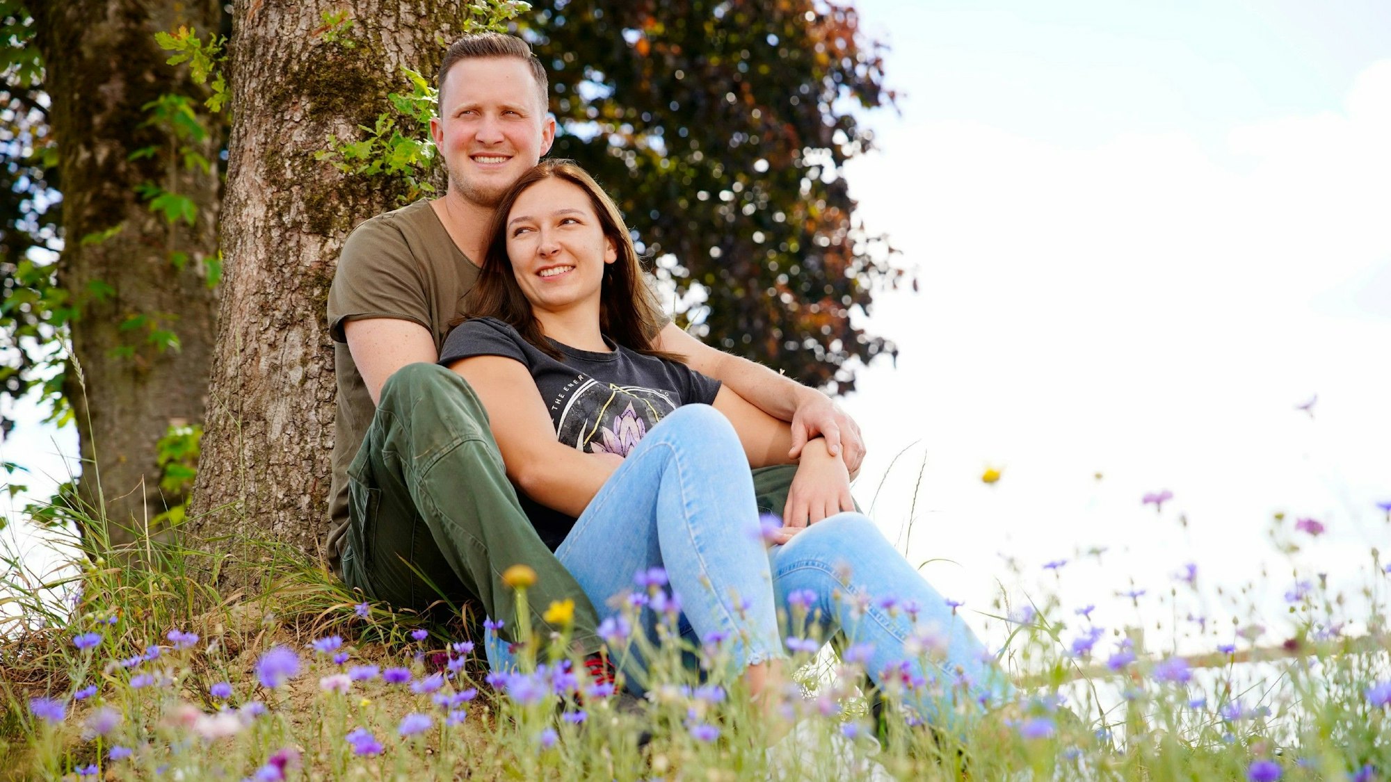 Ein junges Paar sitzt an einen Baum gelehnt auf einer Blumenwiese.