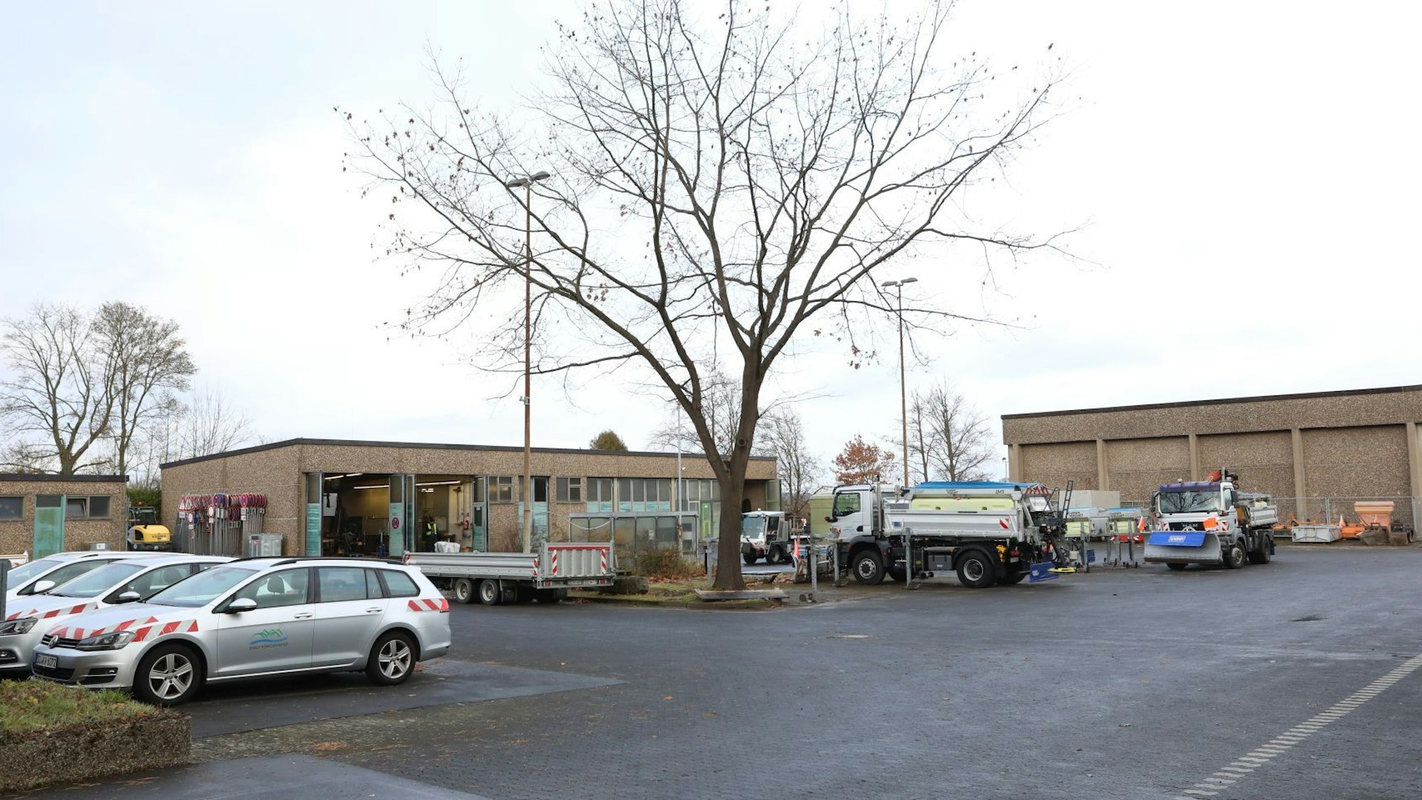 Mehre Fahrzeuge, darunter Lastwagen, stehen auf einer asphaltierten Fläche vor oder neben mehreren Betriebshallen.